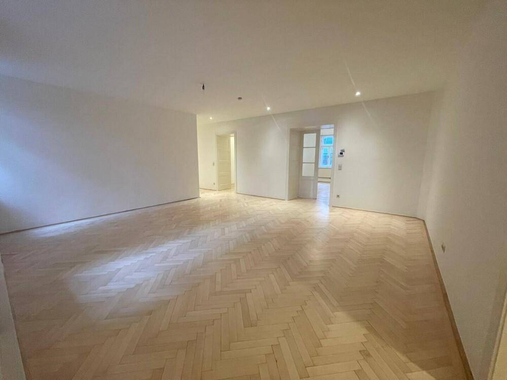 Großer, leerer Wohnraum mit hellem Fischgrätparkett, der zu einem Flur und weiteren Zimmern führt.