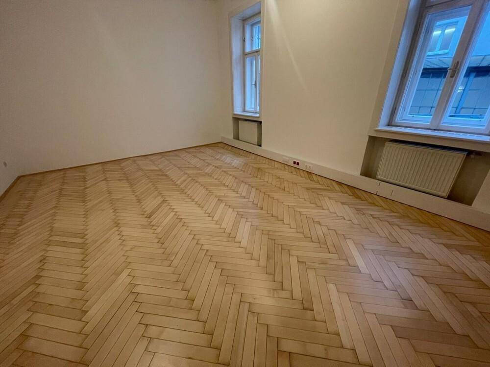 Leerer Raum mit glänzendem Fischgrätparkett und zwei Fenstern, die den Raum mit Tageslicht durchfluten.