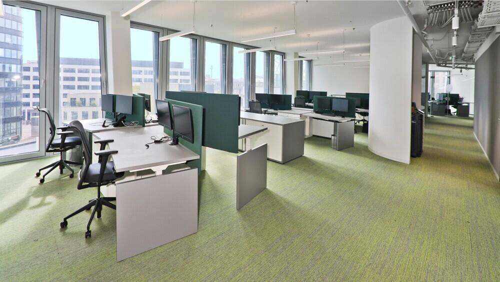 Moderner Großraumbüro mit grünen Teppichfliesen, ergonomischen Arbeitsplätzen und viel Tageslicht durch große Fenster.