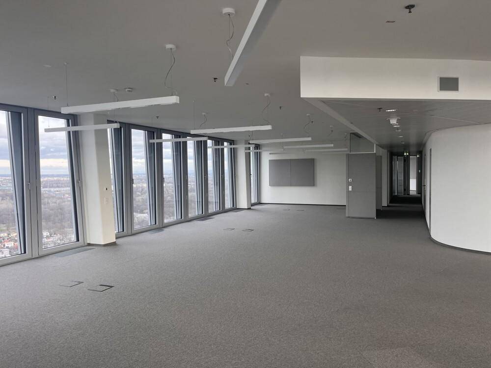 Großer, unmöblierter Bürobereich mit Teppichboden und durchgehenden Fensterfronten, die viel Tageslicht hereinlassen.