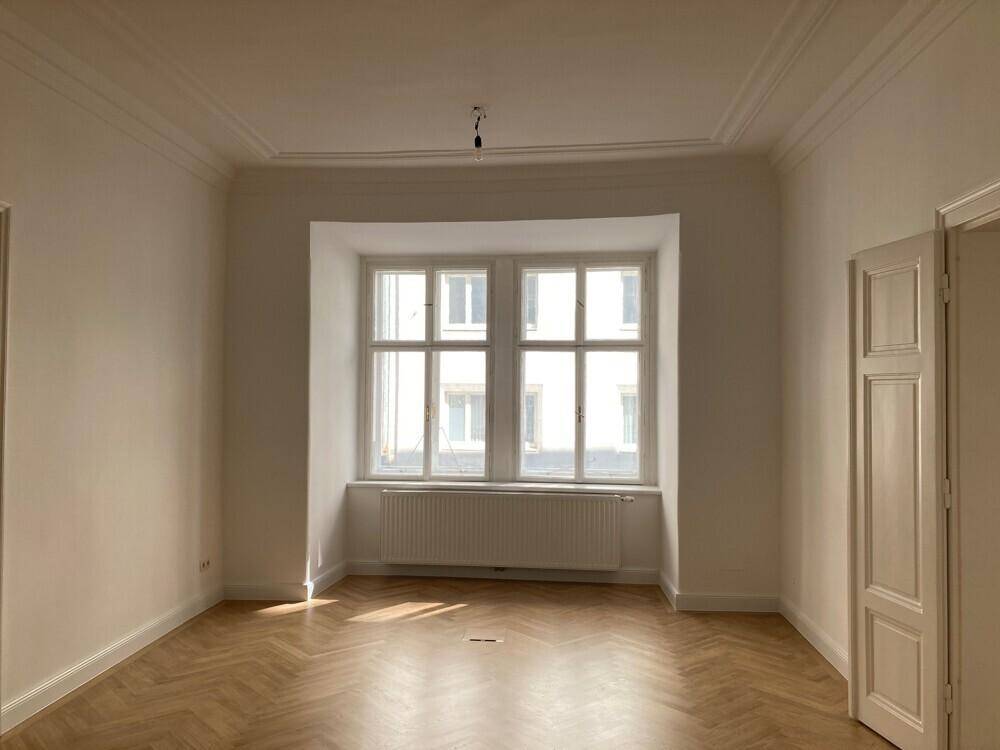 Geräumiges Zimmer mit großem Fenster, Fischgrätparkett und dekorativen Deckenleisten, sehr hell.