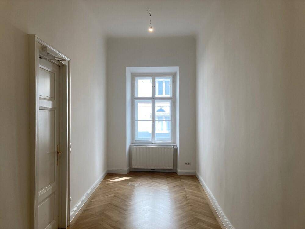 Helles Zimmer mit Fischgrätparkett, einem Fenster und Heizkörper unter der Fensterbank, ideal als Schlafzimmer.