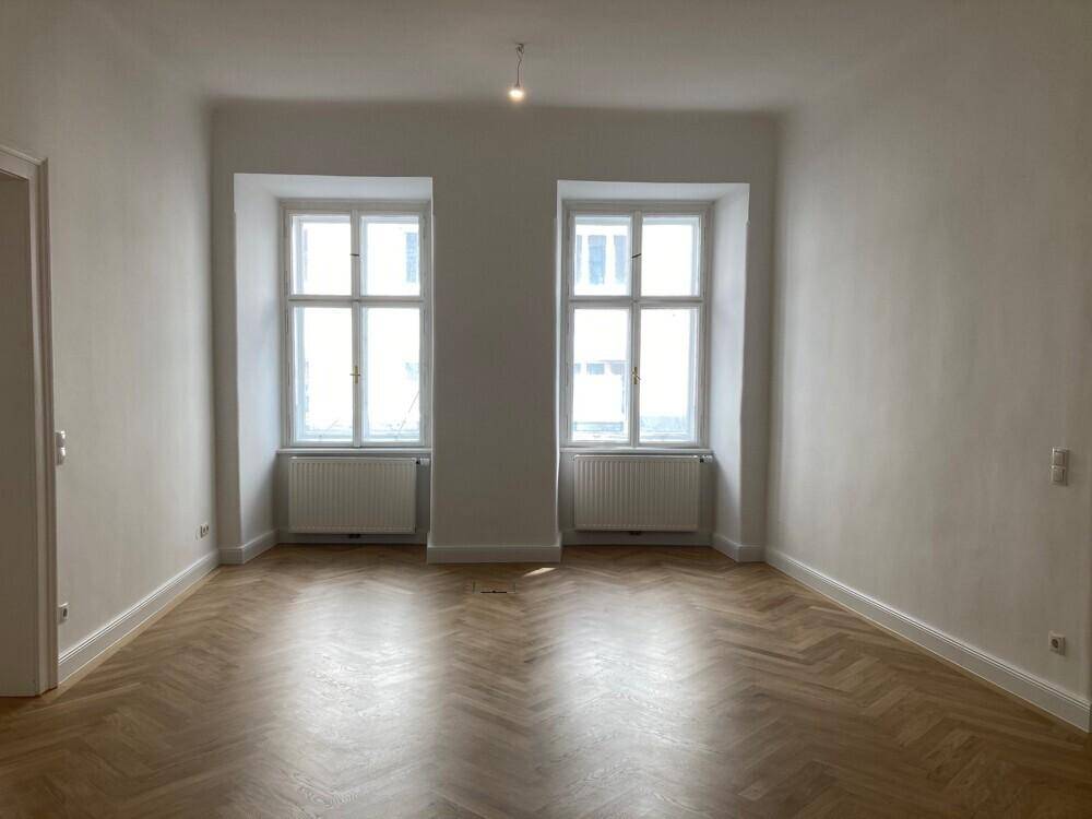 Großer, heller Raum mit zwei Fenstern, Fischgrätparkett und weißen Wänden, bietet viel Tageslicht.