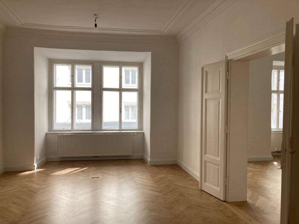 Heller Wohnraum mit großem Fenster, Fischgrätparkett und offener Verbindung zu einem weiteren Zimmer.
