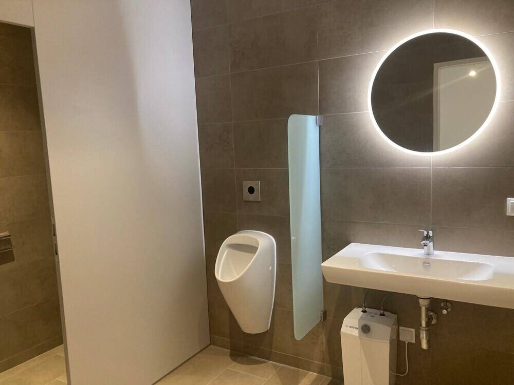 Modernes Badezimmer mit Urinal, Waschbecken und beleuchtetem Spiegel, gefliest in neutralen Tönen.
