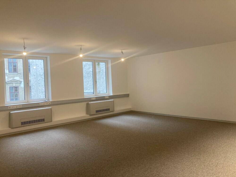 Heller, leerer Büroraum mit Teppichboden und zwei Fenstern, die viel Tageslicht hereinlassen.