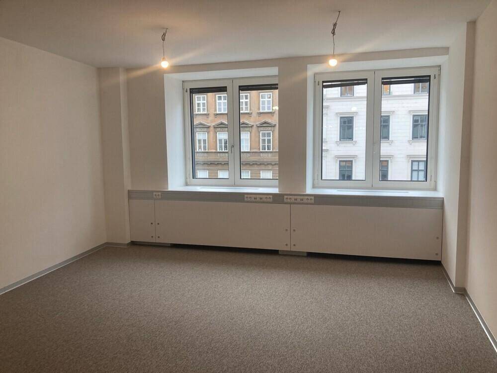 Leerer Büroraum mit zwei großen Fenstern und Teppichboden, ideal für einen Arbeitsplatz.