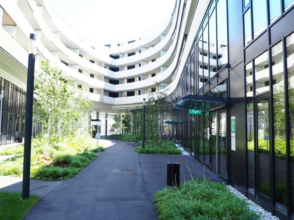 Innenhof eines modernen Gebäudekomplexes mit geschwungener Architektur, viel Grün und einem Fußweg.