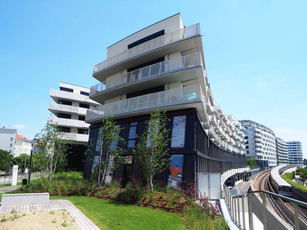 Modernes Wohngebäude mit Balkonen und grüner Landschaftsgestaltung neben Bahngleisen unter blauem Himmel.