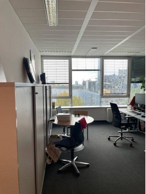 Einzelner Büroraum mit Schreibtischen, Bürostühlen und großen Fenstern mit Stadtblick.