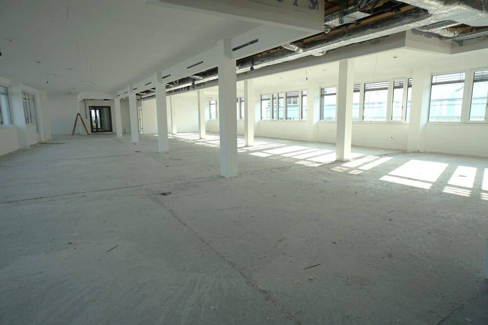 Großer, offener Bürobereich mit Säulen, Rohbetonboden und sichtbaren Deckeninstallationen, viel Tageslicht.