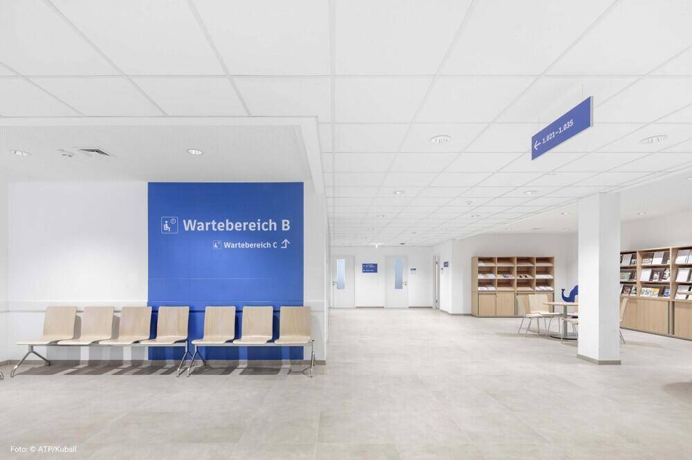 Heller Wartebereich mit bequemen Stühlen, blauer Akzentwand und Informationsregalen in einem modernen Gebäude.