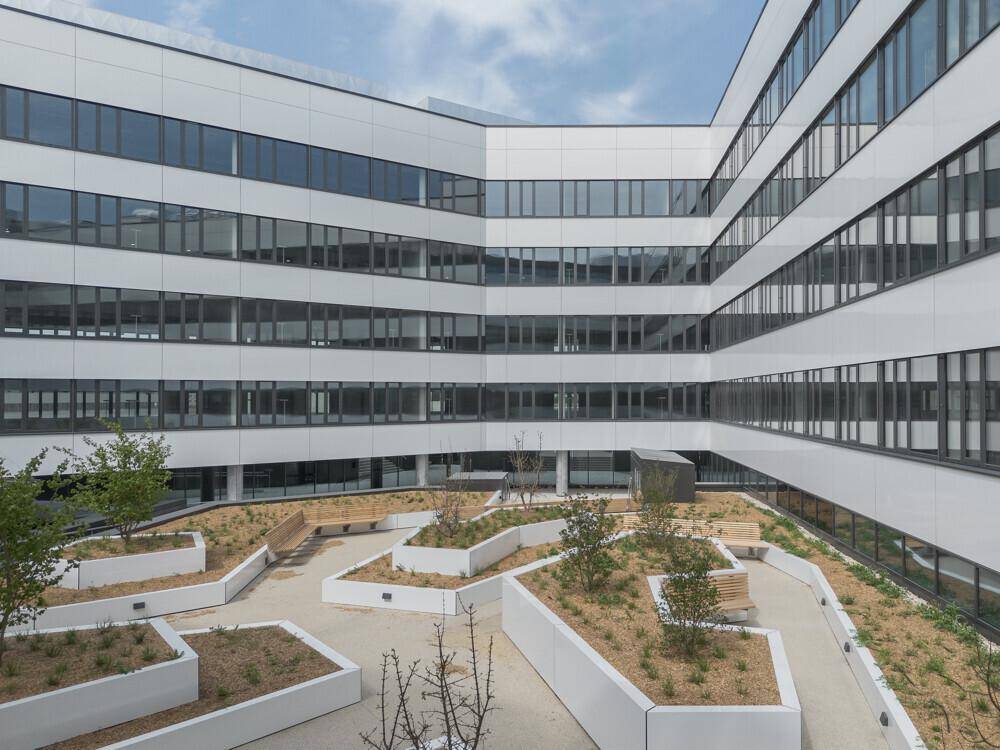 Begrünter Innenhof eines modernen Bürokomplexes mit Sitzgelegenheiten und geometrischen Pflanzbeeten.