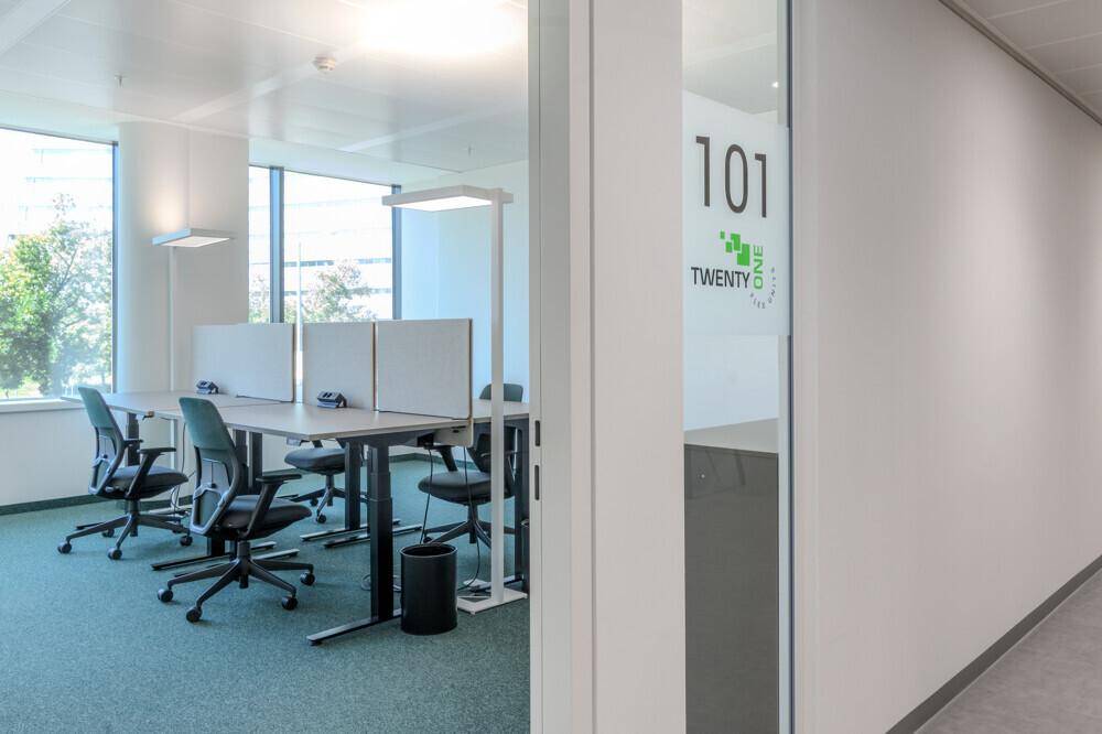 Modernes Büro mit mehreren Schreibtischen, Bürostühlen und großen Fenstern, die viel Tageslicht hereinlassen.