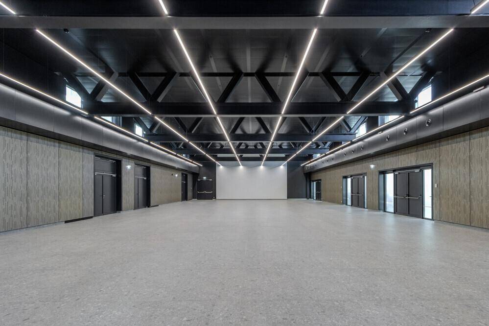 Großer, leerer Saal mit hoher, dunkler Decke, sichtbaren Trägern und modernen Lichtleisten.
