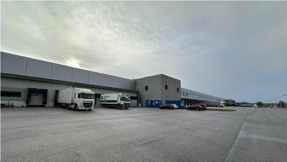 Außenansicht des Logistikzentrums mit mehreren Lastwagen an den Laderampen unter bewölktem Himmel.