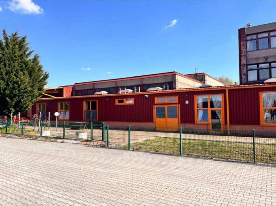 Langes Gebäude mit roter Fassade, vielen Fenstern und einem grünen Zaun im Vordergrund unter blauem Himmel.
