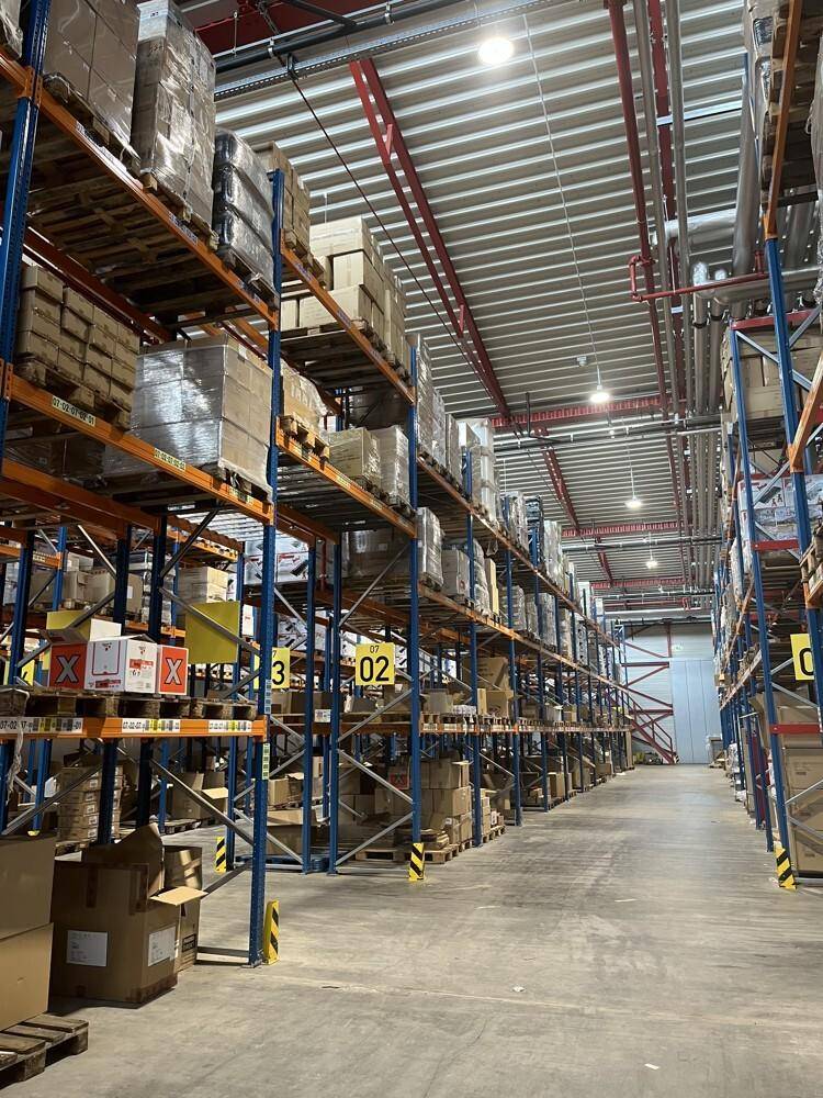 Gang in einer Lagerhalle mit hohen Regalen, die mit verschiedenen Produkten und Verpackungen bestückt sind.