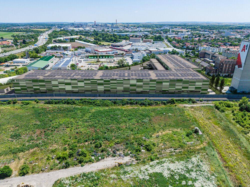 Weitläufige Luftaufnahme eines Industrieareals mit großen Hallen, begrünten Flächen und einer Fernsicht auf die Landschaft.
