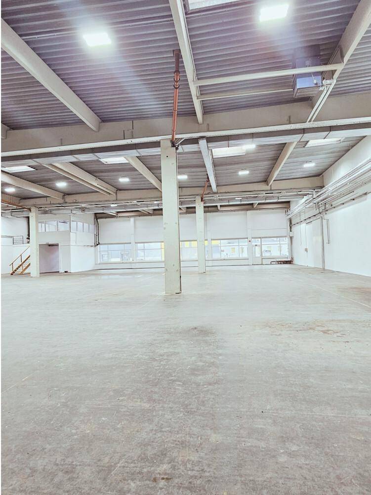 Helle, leere Lagerhalle mit hohen Decken, Betonboden und großen Fenstern, die viel Licht hereinlassen.