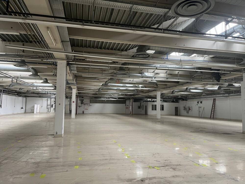 Großer, leerer Gewerberaum mit hohen Decken, sichtbaren Lüftungsrohren und hellen Fliesenböden.