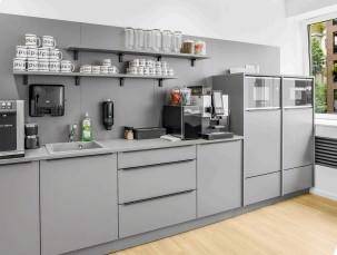 Moderne Teeküche mit grauen Schränken, integrierter Spüle und Kaffeemaschine, ideal für Büroräume.