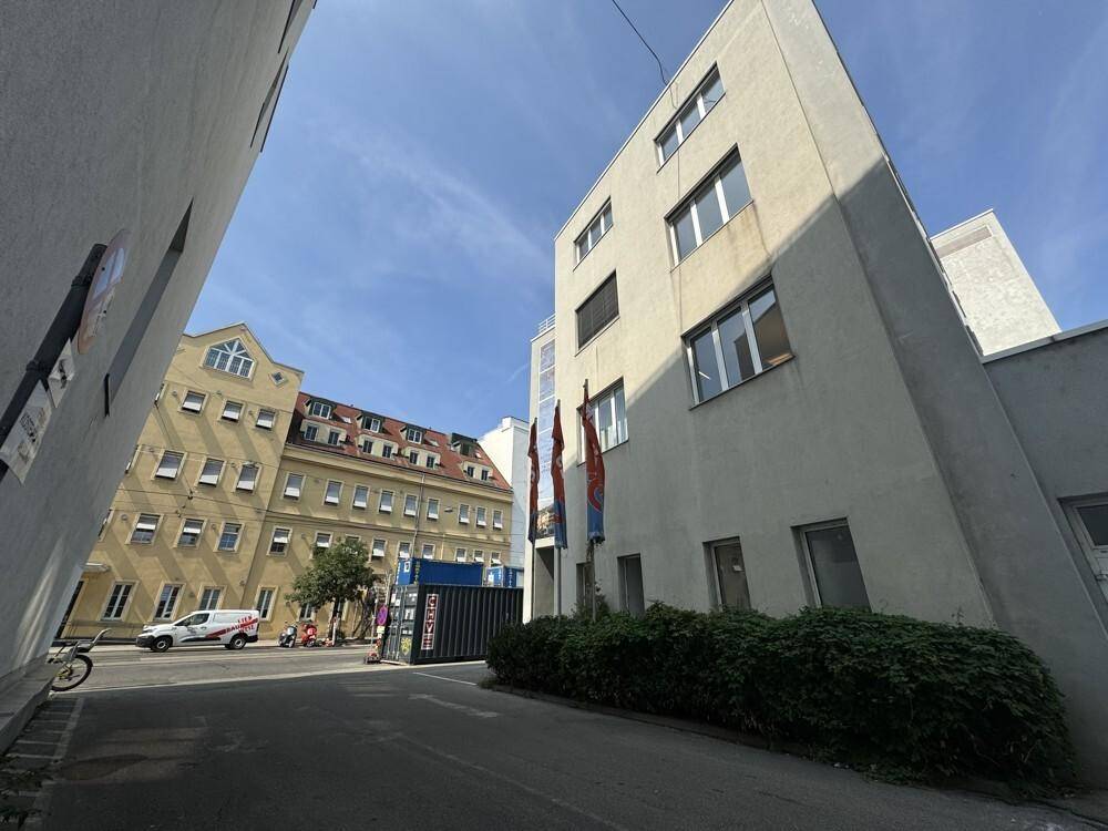 Blick auf zwei Gebäude, eines hellgrau und das andere gelblich, entlang einer schmalen Zufahrtsstraße.