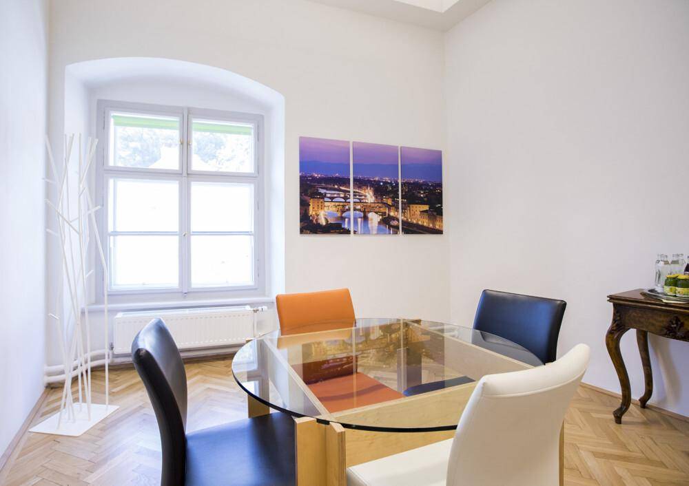 Kleiner Besprechungsraum mit rundem Glastisch, verschiedenen Stühlen und einem Fenster mit Bogen.