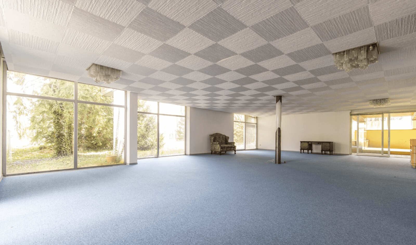 Geräumiger, leerer Raum mit blauem Teppichboden, karierter Decke und großen Fenstern.
