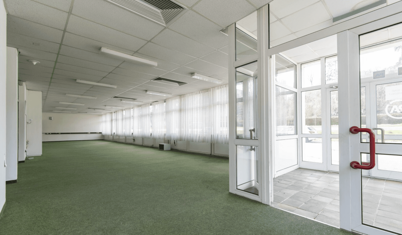 Großer, leerer Raum mit grünem Teppichboden und einer Glasfront am Eingangsbereich.