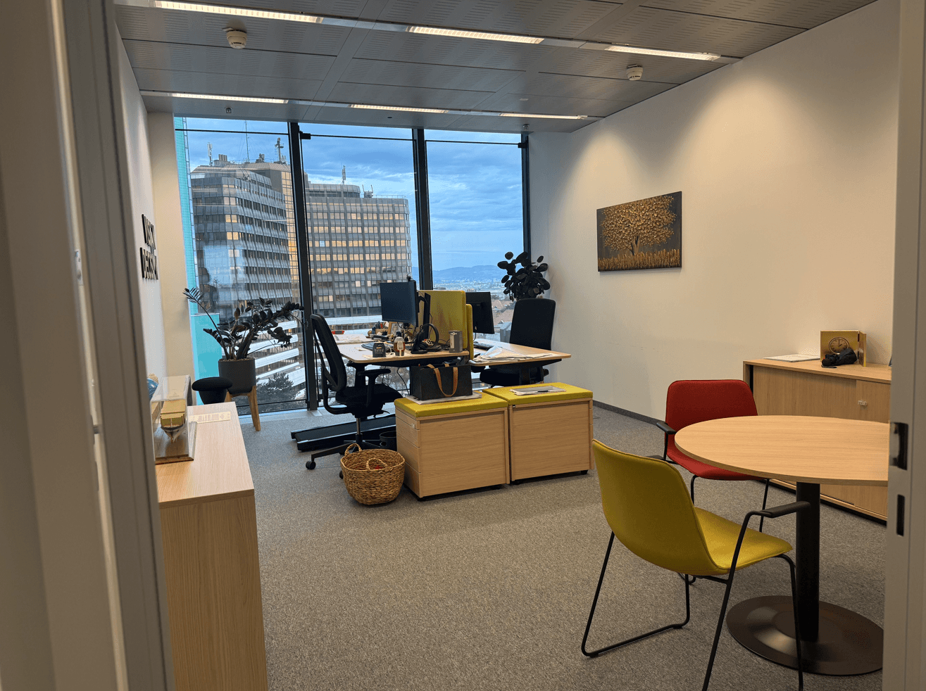 Helles Büro mit großen Fenstern, Blick auf die Stadt und modernen Möbeln in Gelb und Rot.