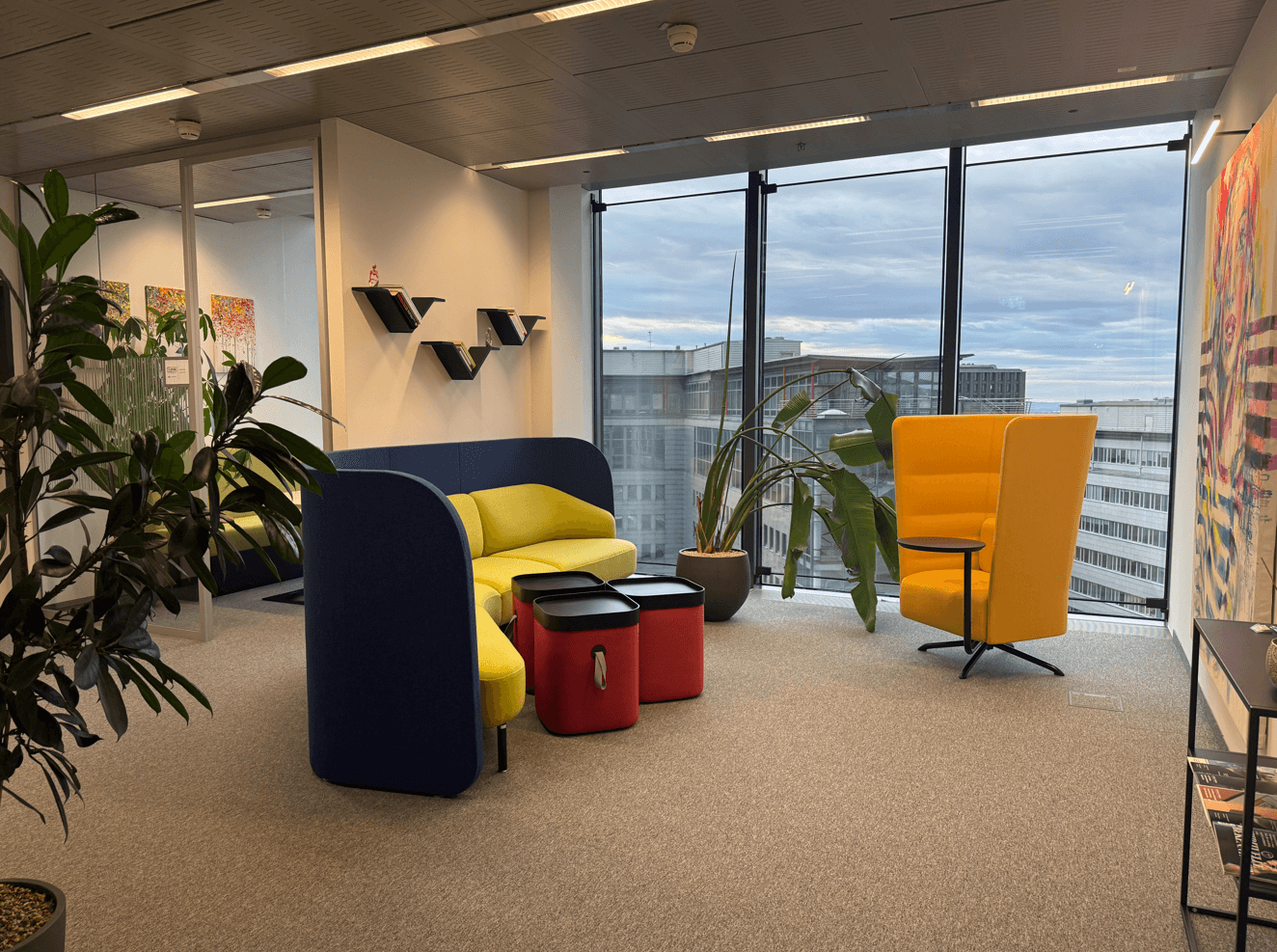 Gemütliche Lounge-Ecke in einem Büro mit farbigen Sitzmöbeln, Pflanzen und großen Fenstern mit Stadtblick.
