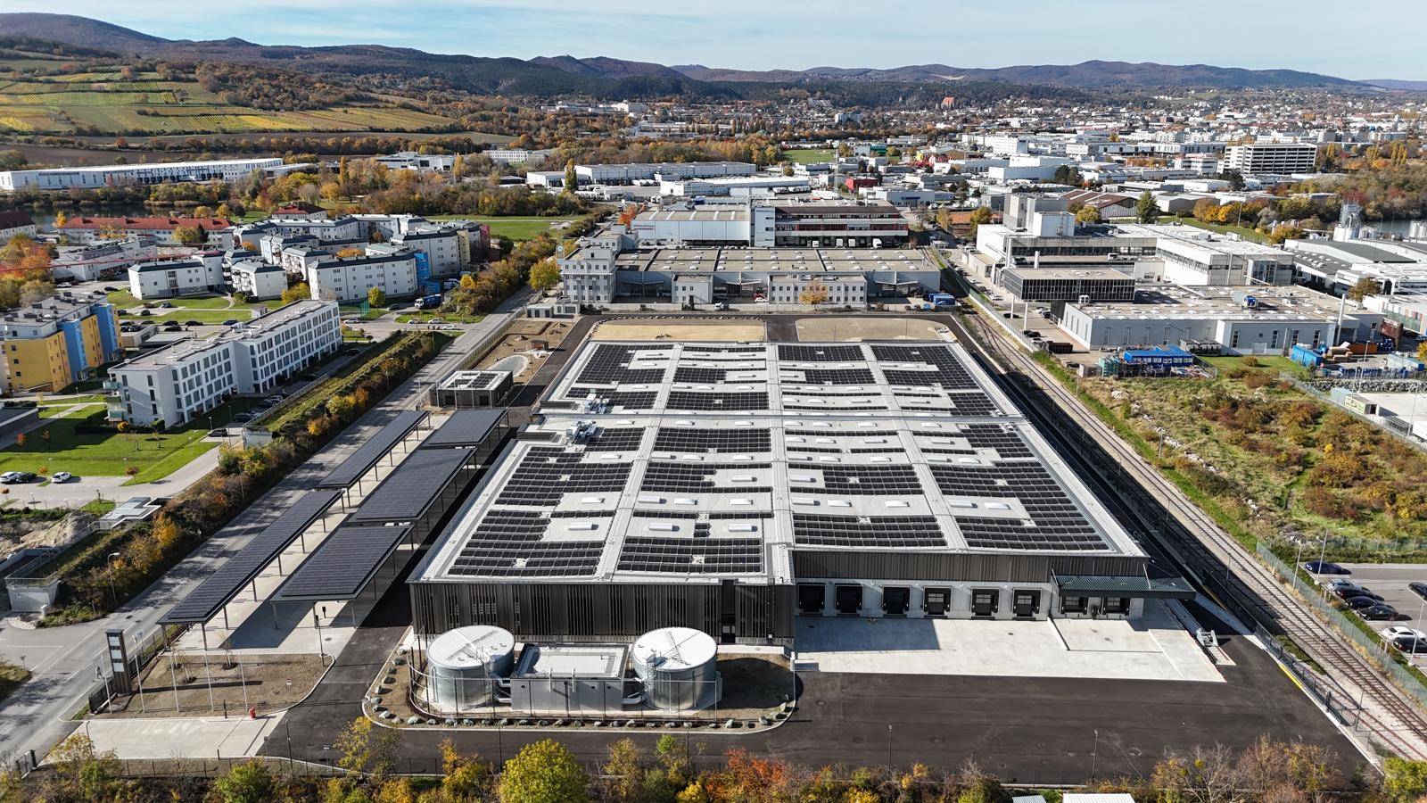 Weitläufige Luftaufnahme eines Industrieareals mit großem Gebäude, Solaranlage und umliegender Bebauung.