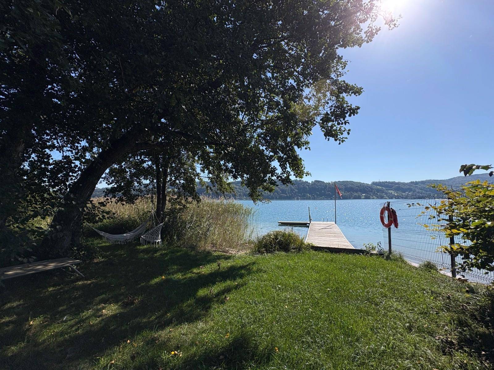 Idyllischer Gartenbereich mit Hängematte und Steg, der in einen klaren See unter strahlendem Sonnenschein führt.