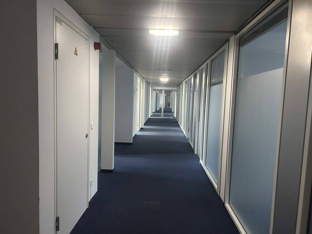 Langer, heller Flur mit blauem Teppichboden und Bürotüren, die zu den Arbeitsbereichen führen.