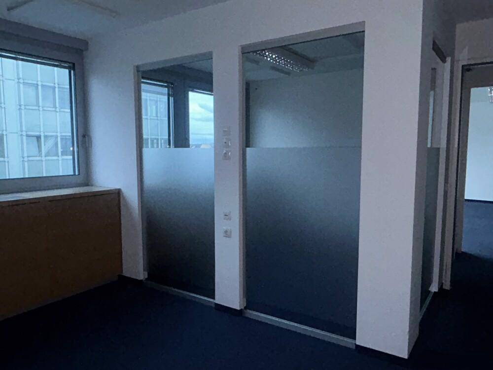 Büroraum mit blauem Teppichboden, Fenster und Milchglastüren für Privatsphäre.