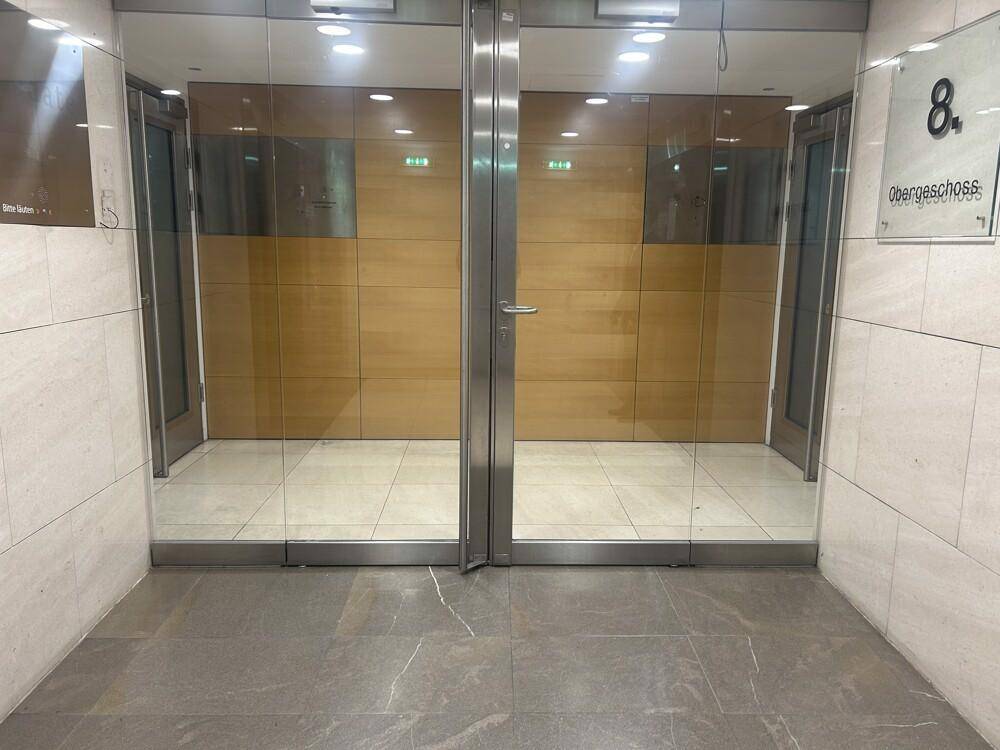 Aufzugsbereich mit Glastüren und Fliesenboden, der zu den oberen Stockwerken führt.