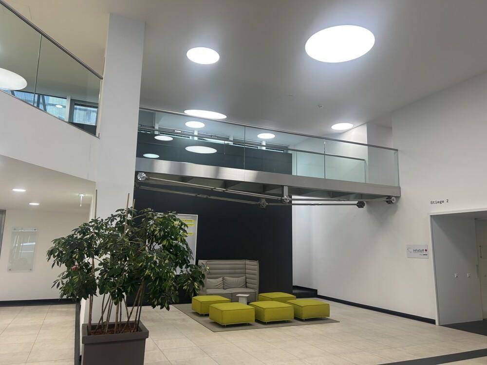 Geräumige Lobby mit Sitzgelegenheiten, Pflanzen und Blick auf eine obere Etage mit Glasgeländer.
