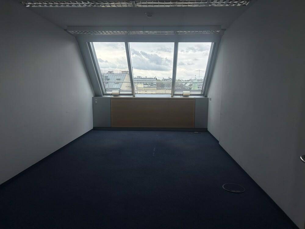 Büroraum im Dachgeschoss mit blauem Teppichboden und schrägen Fenstern mit Stadtblick.