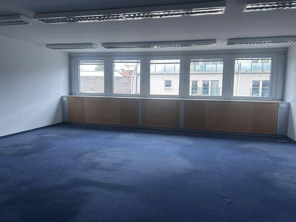 Leerer Büroraum mit blauem Teppichboden und einer Reihe von Fenstern, die viel Licht spenden.