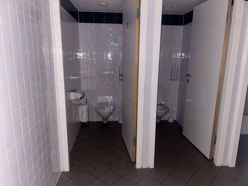Zwei Toilettenkabinen mit weißen Fliesen und dunklem Boden in einem öffentlichen Waschraum.