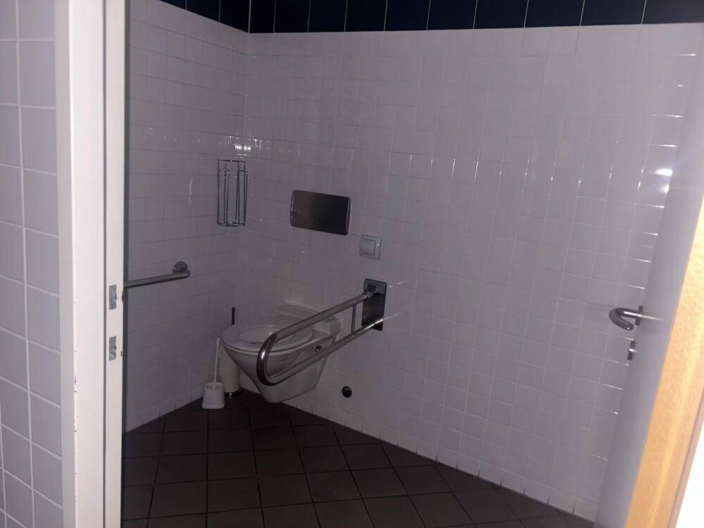 Barrierefreie Toilette mit Haltegriffen, weißen Fliesen und dunklem Boden für Rollstuhlfahrer.