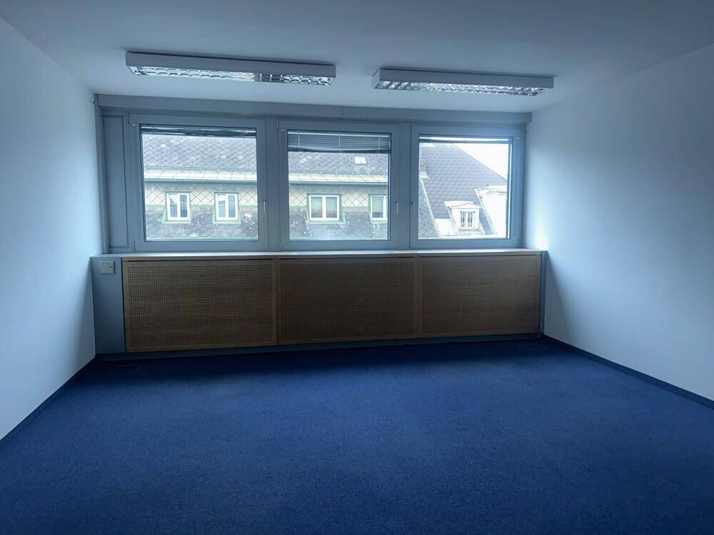 Leerer Büroraum mit blauem Teppichboden und Fenstern, die viel Tageslicht hereinlassen.