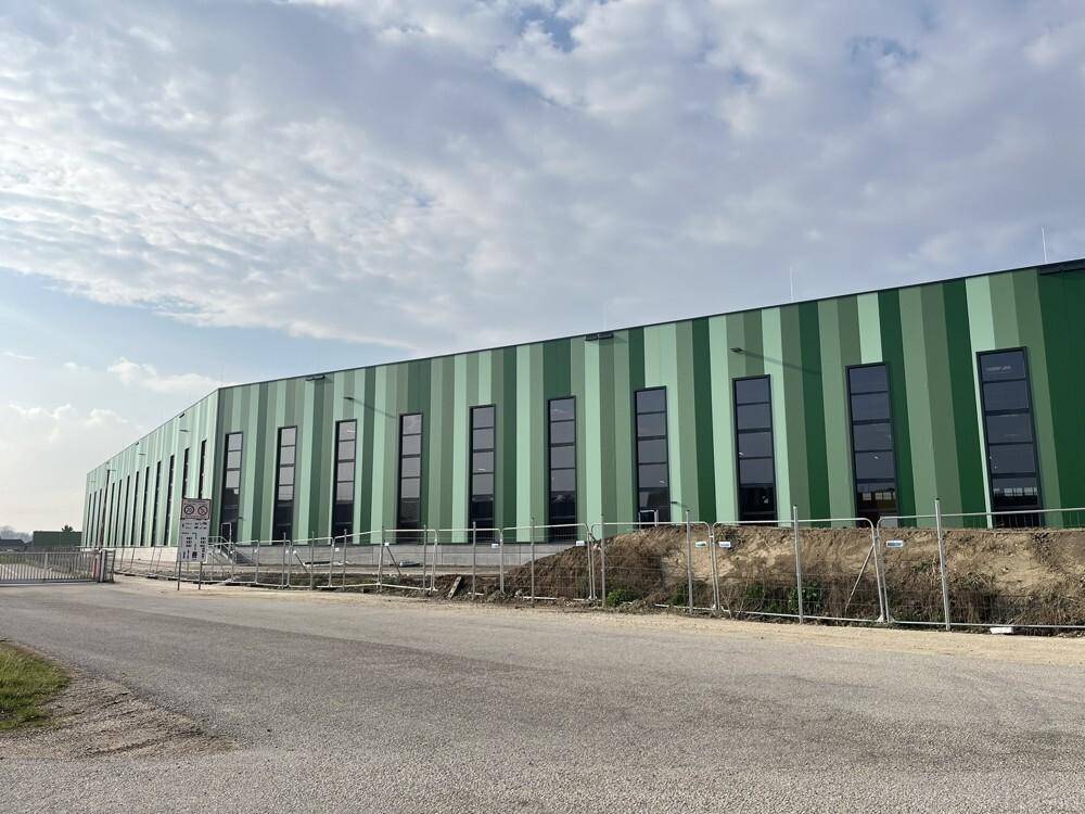 Die lange, gestreifte grüne Fassade des Industriegebäudes mit vertikalen Fenstern und einem Zaun im Vordergrund.
