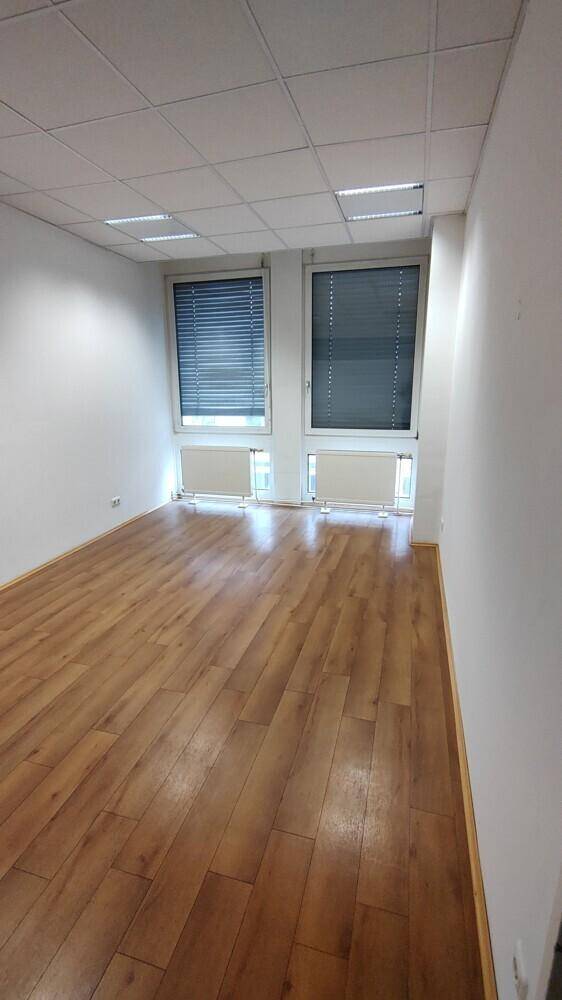 Heller Büroraum mit Holzboden, Rasterdecke und zwei Fenstern mit Jalousien für optimale Lichtregulierung.