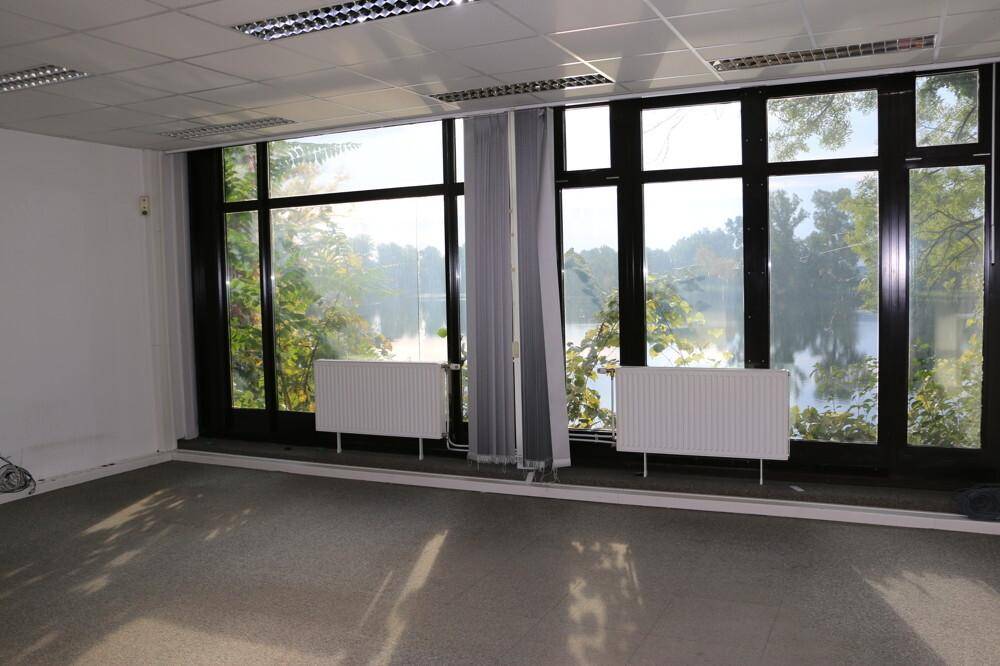 Heller Büroraum mit großen Fenstern, Blick ins Grüne und auf Wasser, ausgestattet mit Terrazzo-Boden und Heizkörpern.