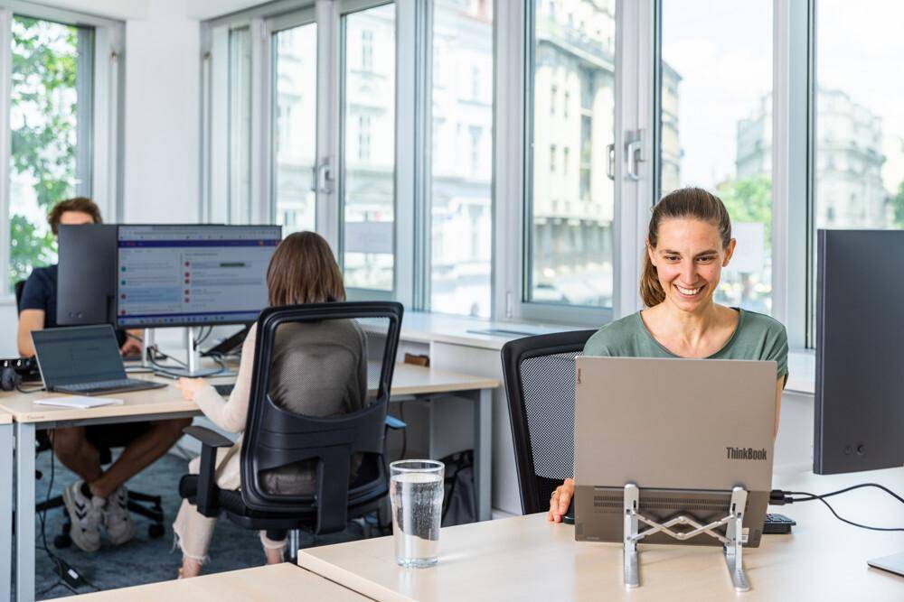 Mitarbeiter arbeiten an ihren Schreibtischen in einem hellen Büro mit großen Fenstern und Stadtblick.