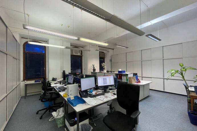 Großraumbüro mit mehreren Arbeitsplätzen, grauen Teppichböden und modernen Deckenleuchten.