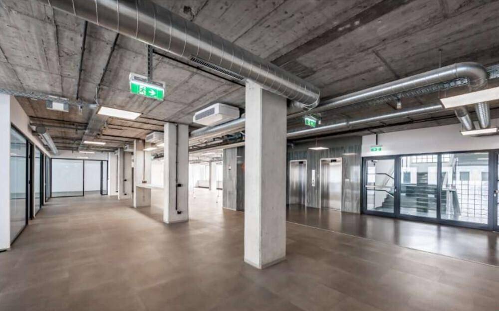 Offener Flur mit Betonstützen, freiliegenden Rohren und Blick auf Aufzüge und Treppenhaus.