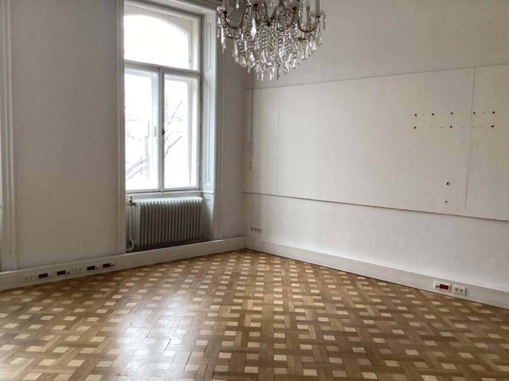 Helles Zimmer mit Parkettboden, Kronleuchter und großen Fenstern, die viel Licht hereinlassen.
